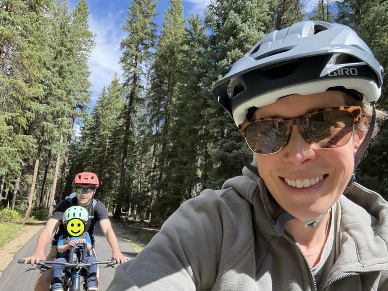The founder smiling on a paved forest path, with her husband and their son, in a front-mounted child seat, riding right behind her; a smiley face covers the son's face