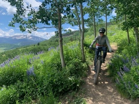 The founder mountain biking down a narrow dirt trail through aspens and purple wildflowers, with snowy peaks behind